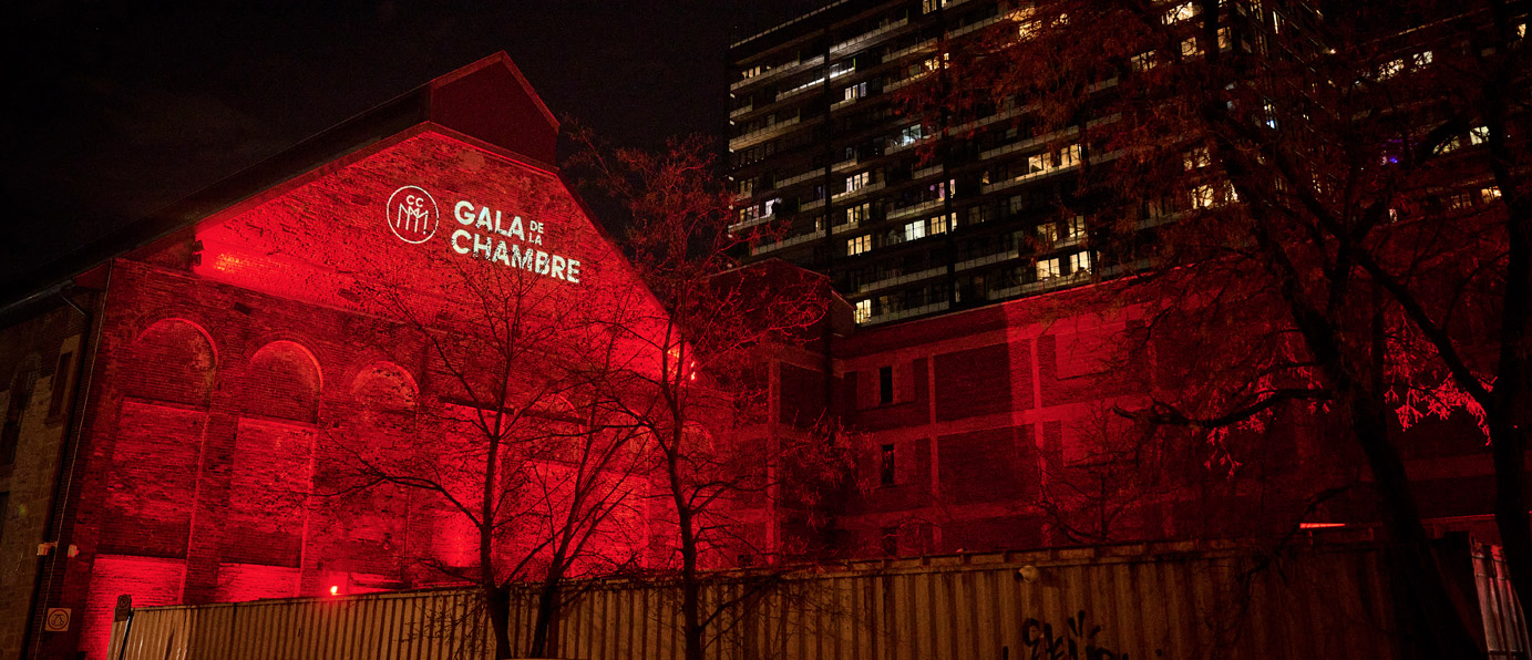 A building lit with striking red lighting at night, featuring the text Gala de la Chambre projected on its façade, surrounded by urban scenery.