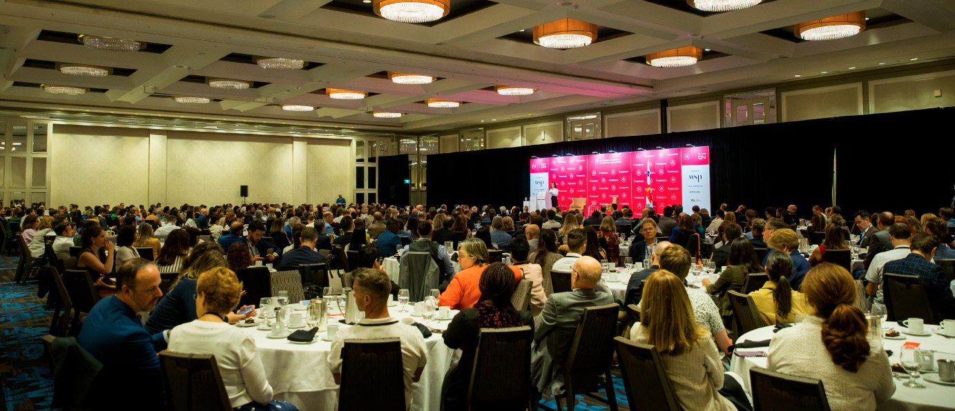 A large formal event with numerous people seated at tables in a ballroom, facing a stage with red branding and speakers presenting